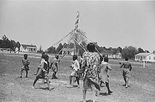 African-American children dancing around a pole holding ribbons tied to the top.
