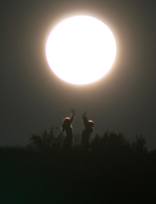 Two people standing facing each other reaching toward large overexposed full moon.