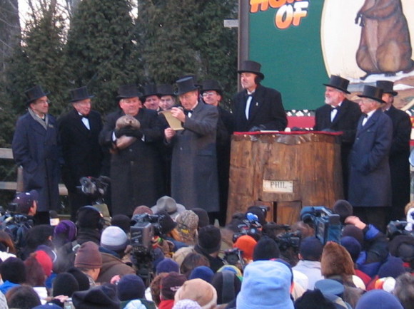 Men in dark winter coats and hats, one holding a groundhog; crowd watching.