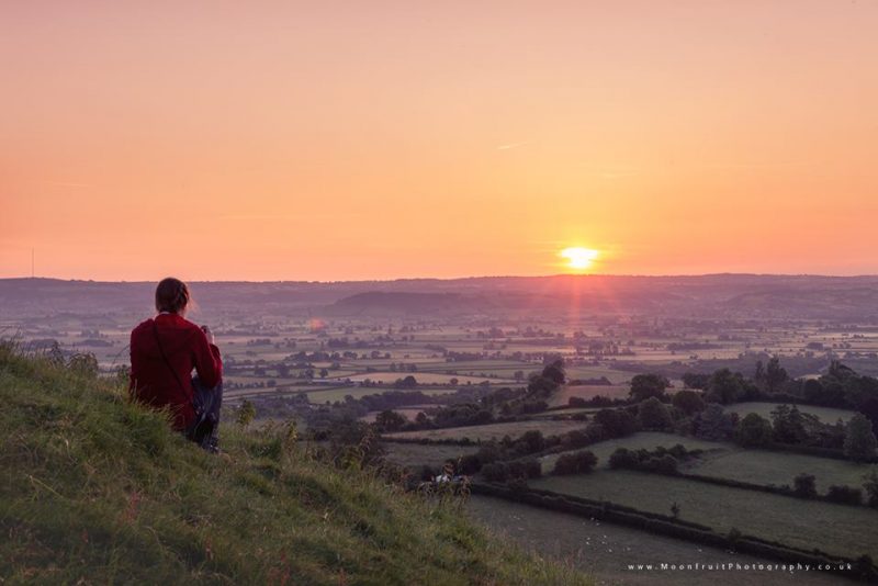 Man sitting on hillside with wide, distant rolling landscape, sun near horizon.