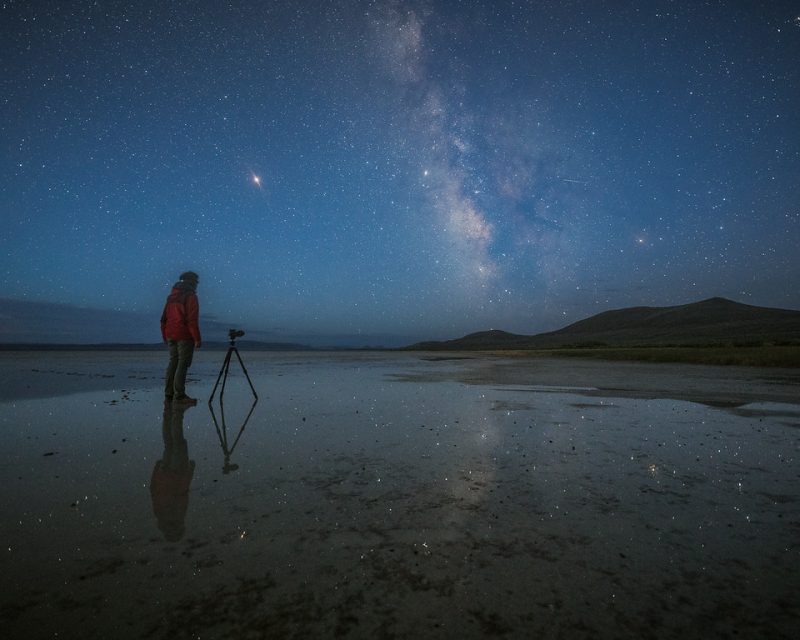 Man with camera on tripod, rising Milky Way reflected on flat water-covered ground.