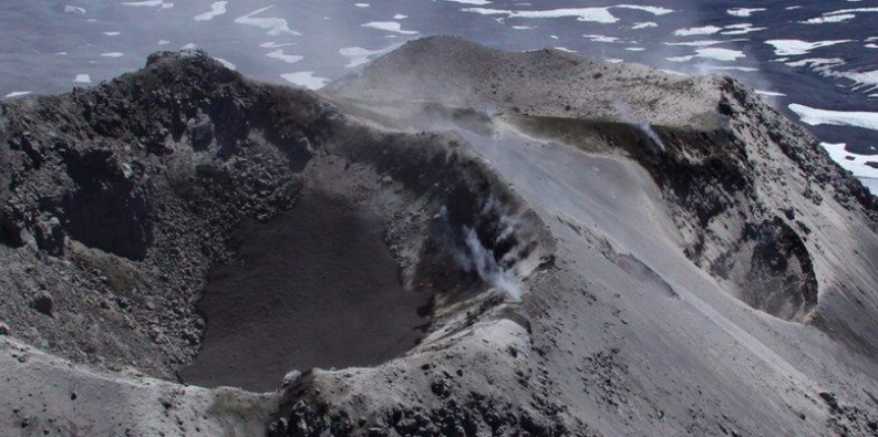 Aerial view of crater with water in it on an ash-covered mountain top.