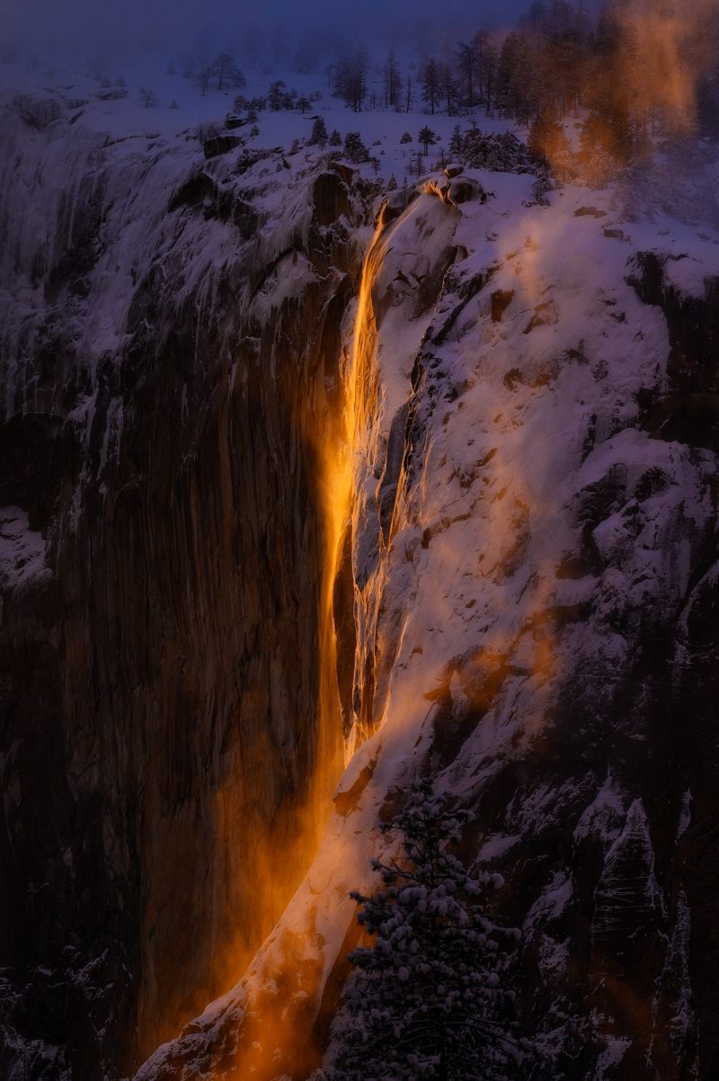 Tall, thin waterfall on rocky cliff lit by an orange glow.