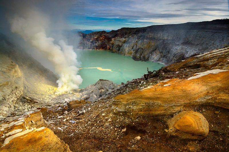 Steaming greenish lake in deep volcanic crater with yellow sulfur deposits on shore.