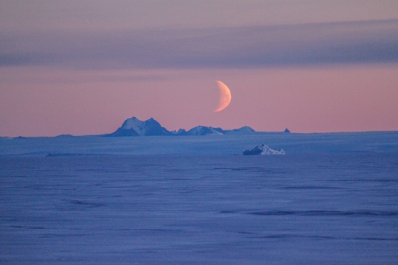 Icy landscape to a distant horizon, with ice hills and orange crescent moon above.