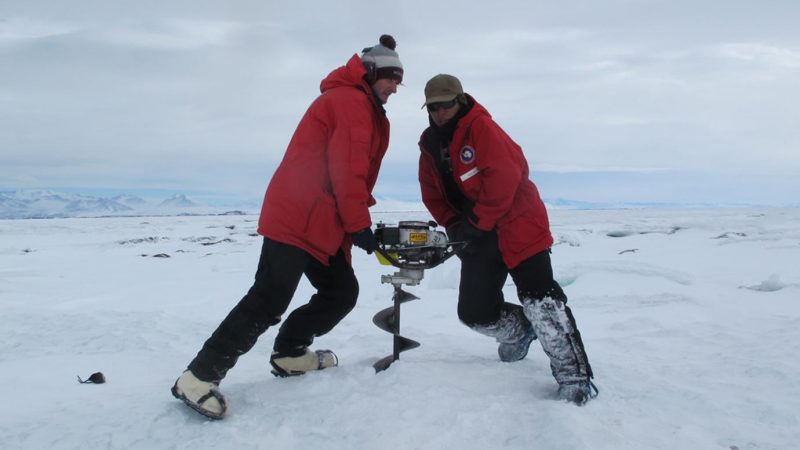 Two heavily-clothed scientists straining to twist a large auger into the ice.