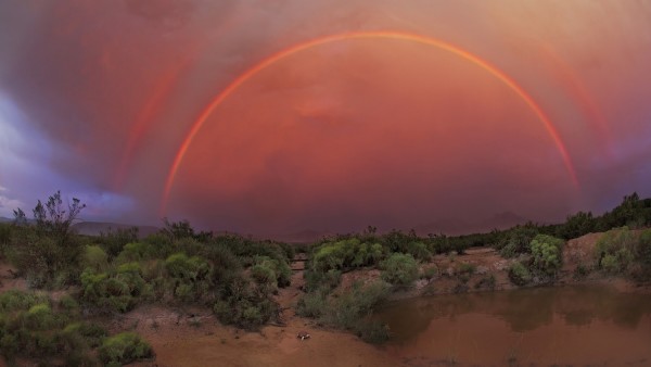 Two concentric red semicircular arcs against deep orange clouds over a brushy desert lanscape.