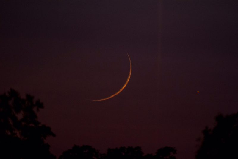 Very thin orange crescent in deep reddish purple sky with a bright dot to its right.