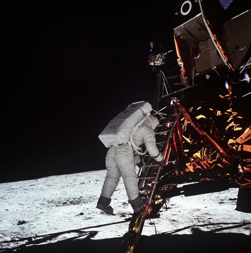 Astronaut on ladder from gold foil covered lunar module, moon horizon and black sky in background.
