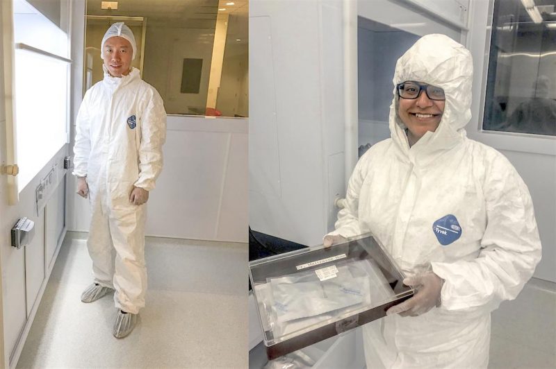 2 grinning scientists in white protective suits, one holding a glass-topped sample box.