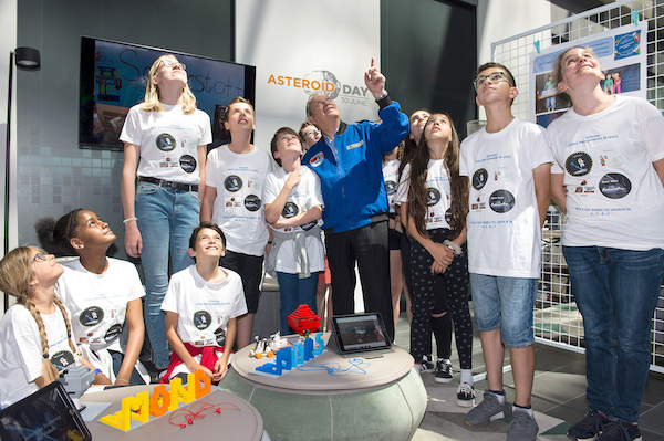 Bunch of children and teens in logo t-shirts looking up in a tall well-lit room.