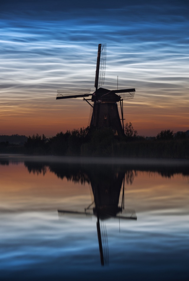 Shining blue clouds at night. Silhouette of Dutch windmill. Reflections in water.