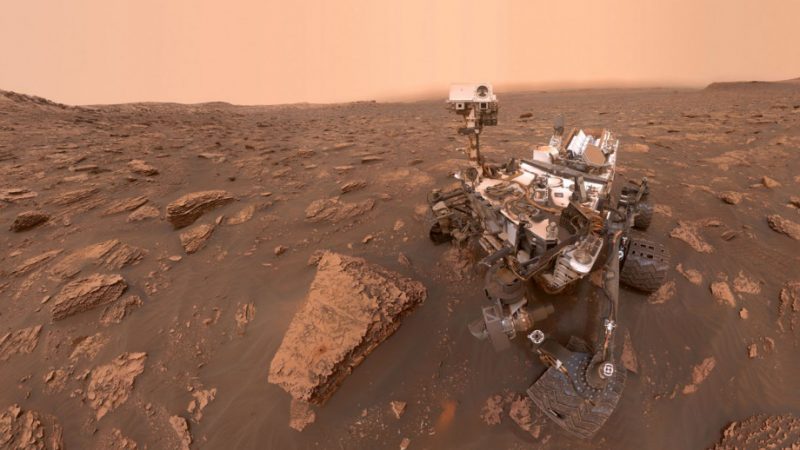Complicated machine on wheels in a red-brown rocky landscape under a pale orange sky.