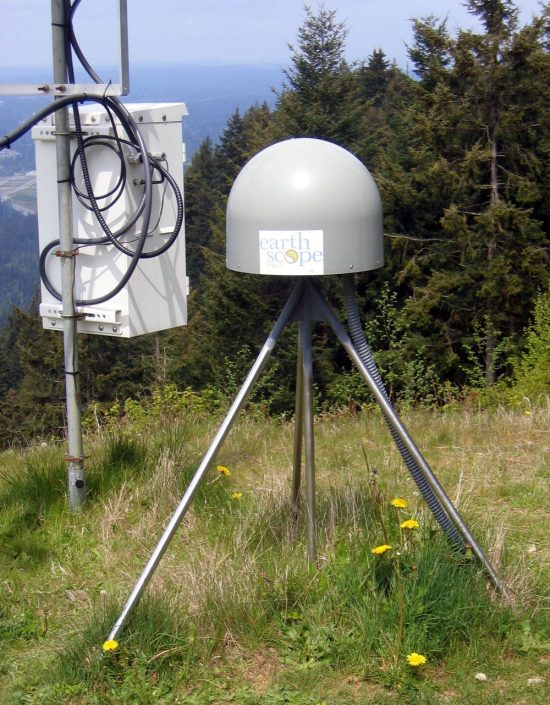 Metal dome on tripod of three short poles next to a metal box on a pole, in a field.