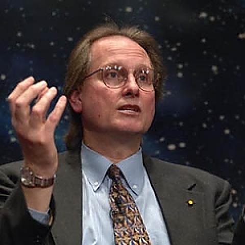 Man in suit gesturing with one hand against out-of-focus starry background.