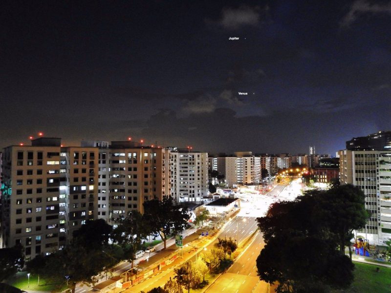 Vênus e Júpiter em uma rua iluminada da cidade, com prédios de vários andares e luzes de carros.