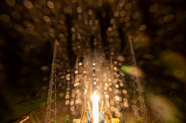Rocket launch from low angle with flame beneath rocket and many bright spots in foreground.