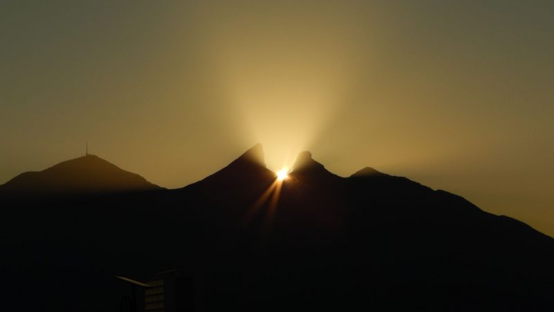 Mountain with yellow-orange light emanating between 2 peaks and sun visible between them.