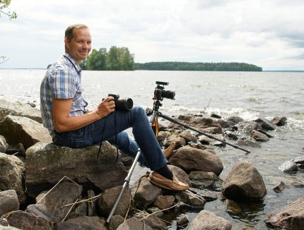 A man with a camera and tripod, seated by a body of water.