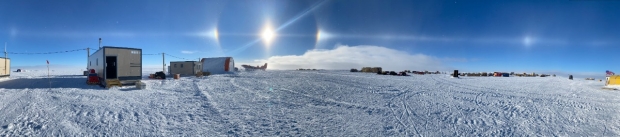 Solar halo and sundogs over Antarctic research camp on vast snow field.